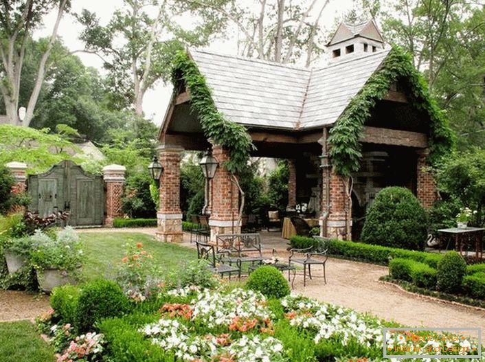 Simplitatea elegantă a unui foișor deschis în stilul unei cabane arată organic într-o grădină de lux și confortabilă. 